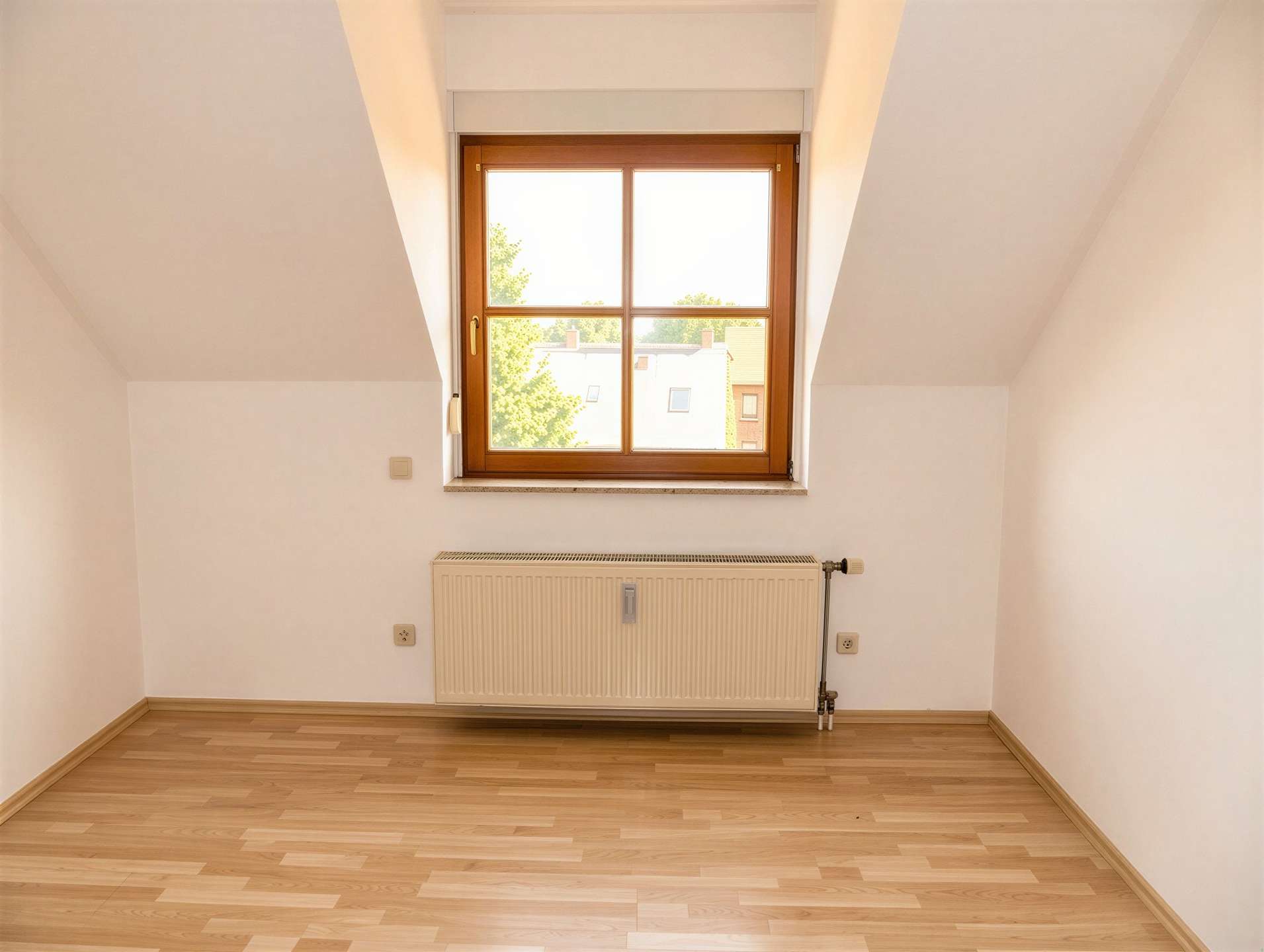 Empty attic room with a wooden-framed window, radiator beneath, and light hardwood flooring.