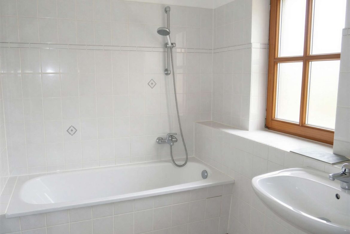 White tiled bathroom featuring a built-in tub with a wall-mounted shower and a handheld shower head, and a wooden-framed window to the right