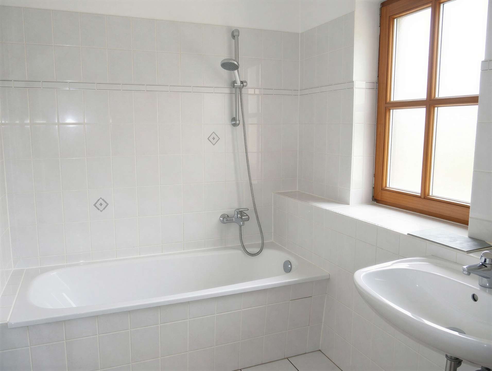 White tiled bathroom featuring a built-in tub with a wall-mounted shower and a handheld shower head, and a wooden-framed window to the right