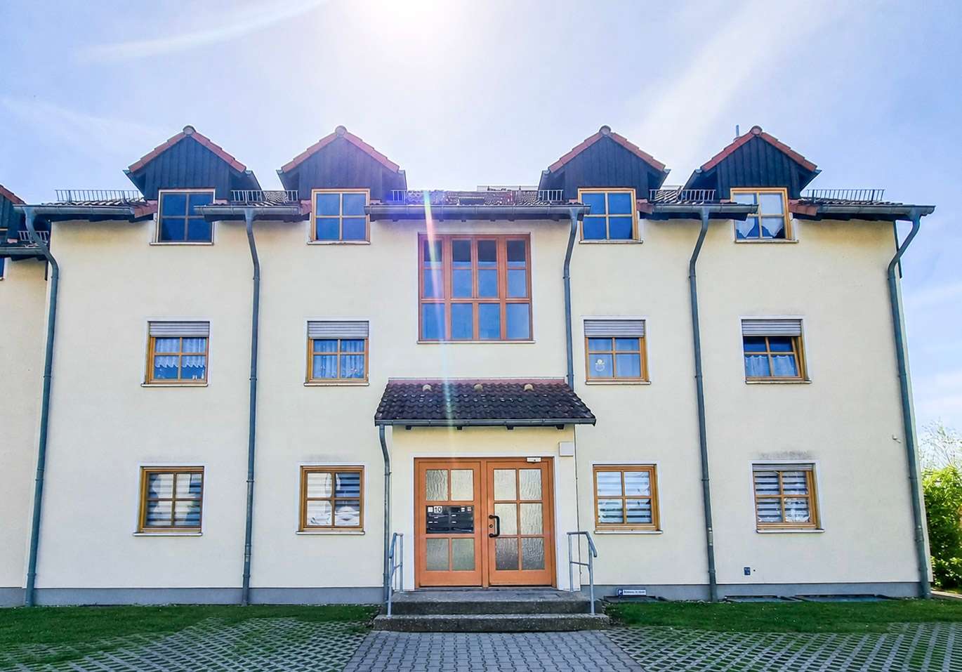 Cream-colored residential building with orange-framed windows, central glass double doors, and a small tiled porch under a sunny sky.