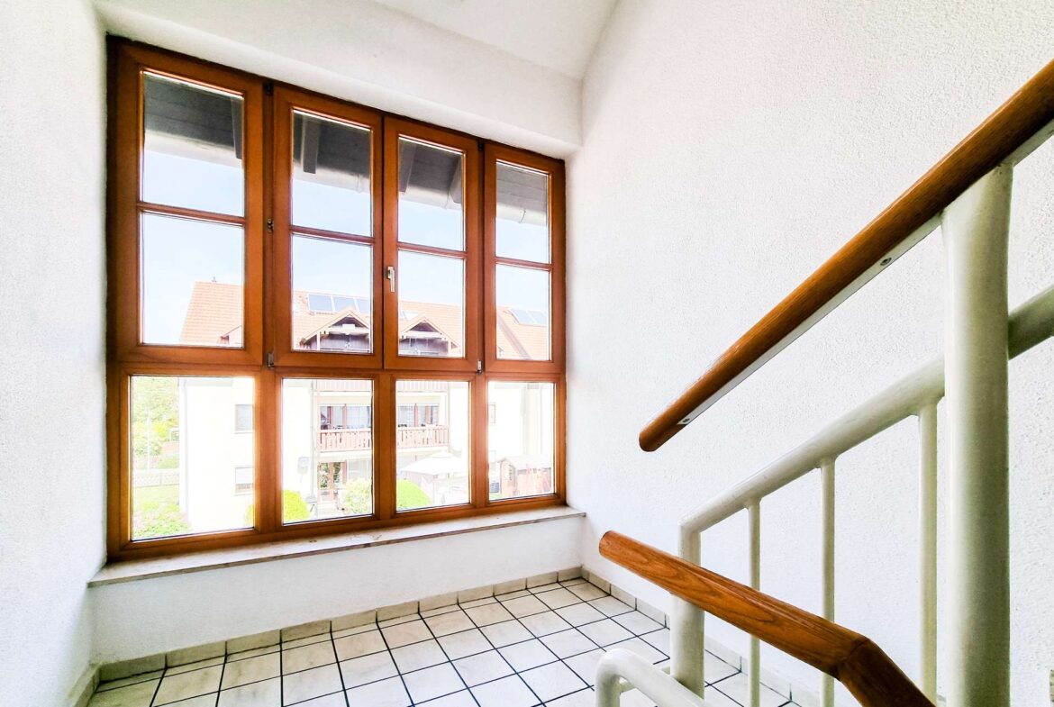 Indoor stairwell with large wooden-framed windows, white walls, and a brown handrail; tiled floor visible at the bottom of the stairs.