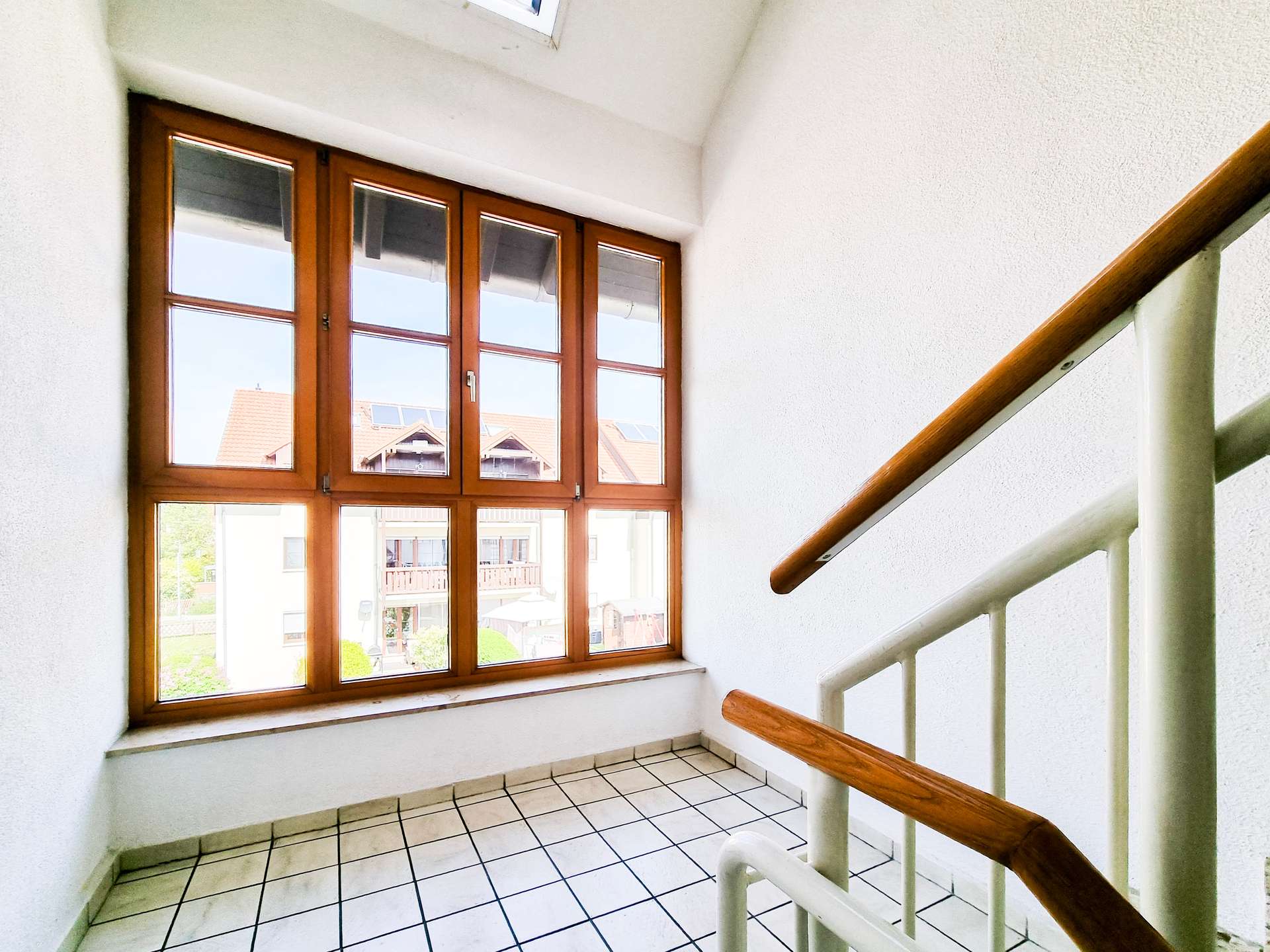 Indoor stairwell with large wooden-framed windows, white walls, and a brown handrail; tiled floor visible at the bottom of the stairs.