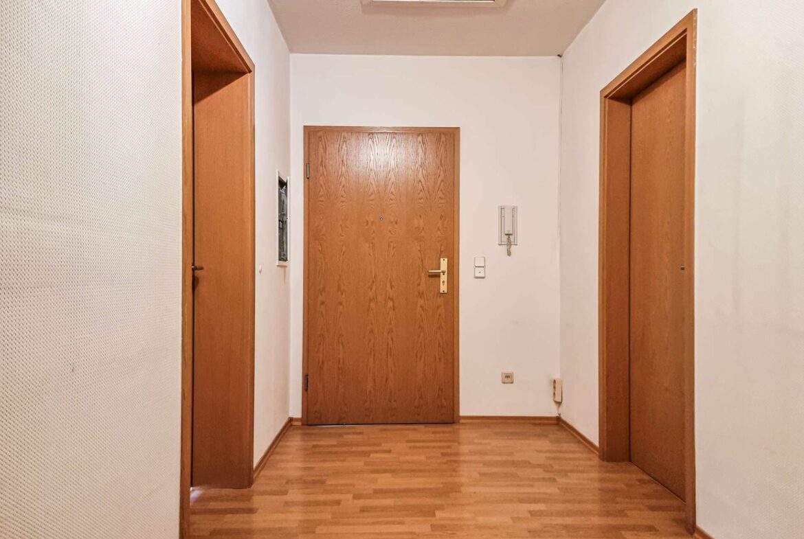 Long hallway with three wooden doors, central door at end, and wooden floor; intercom and light switch on the right wall.
