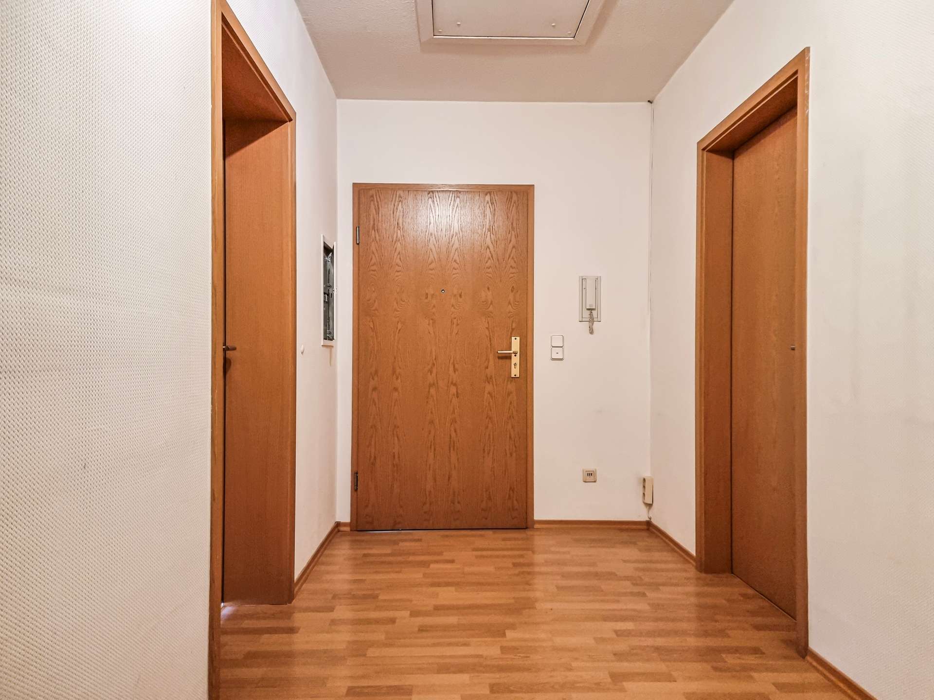 Long hallway with three wooden doors, central door at end, and wooden floor; intercom and light switch on the right wall.