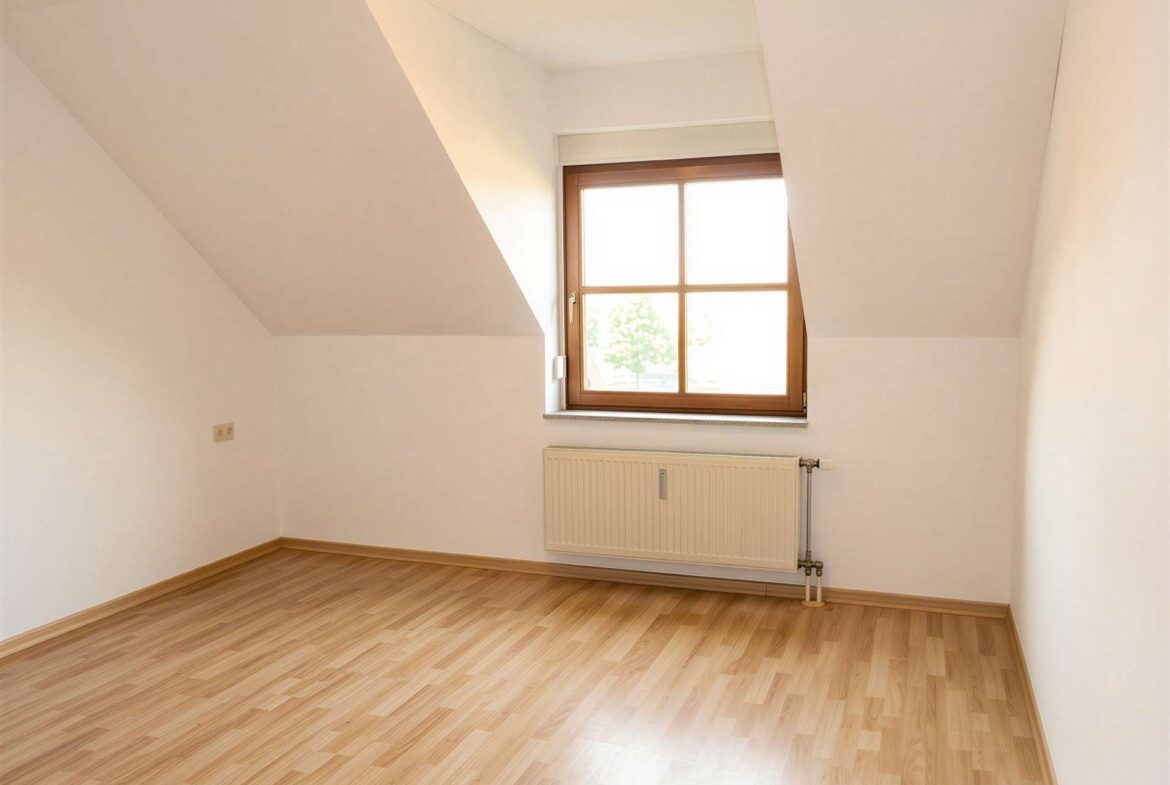 Empty attic room with slanted white walls, wooden floor, and a window letting in bright natural light, with a radiator beneath it.