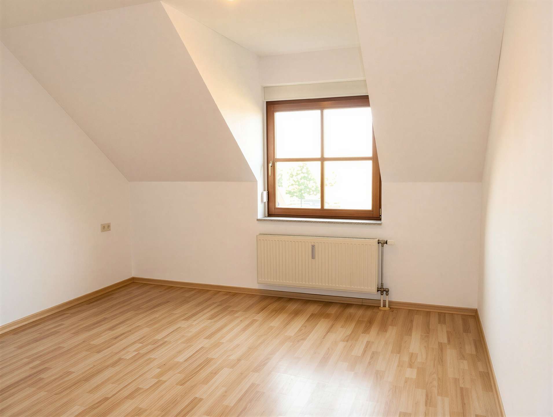 Empty attic room with slanted white walls, wooden floor, and a window letting in bright natural light, with a radiator beneath it.
