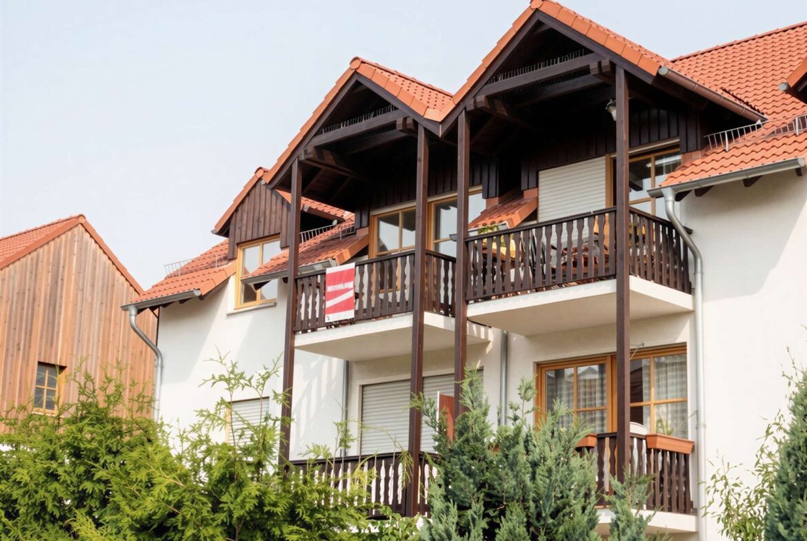 Three-story apartment building with dark wood framing, red tile roof, and multiple balconies, partly obscured by green shrubs in the foreground.
