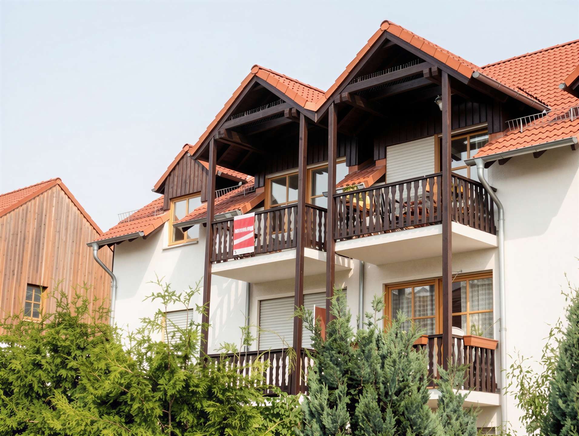Three-story apartment building with dark wood framing, red tile roof, and multiple balconies, partly obscured by green shrubs in the foreground.