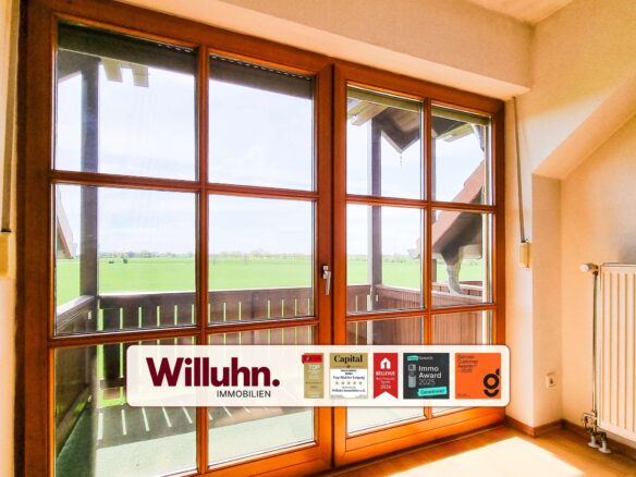 Interior view of a room with large wooden-framed glass doors looking out onto green fields; a Willuhn Immobilien sign with awards is visible on the glass.