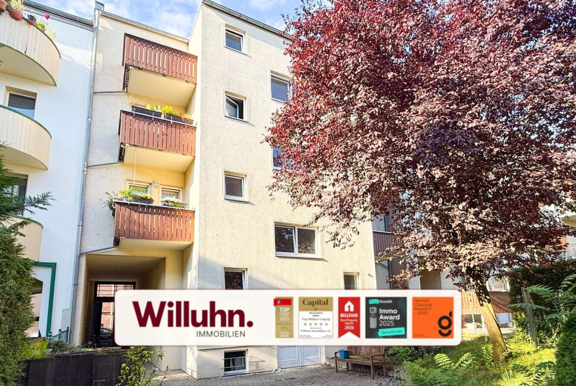 Apartment building with brown balconies, a large red-leaved tree on the right, and a Willuhn Immobilien sign with awards at the entrance.