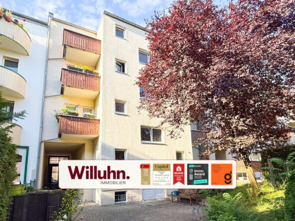 Apartment building with brown balconies, a large red-leaved tree on the right, and a Willuhn Immobilien sign with awards at the entrance.