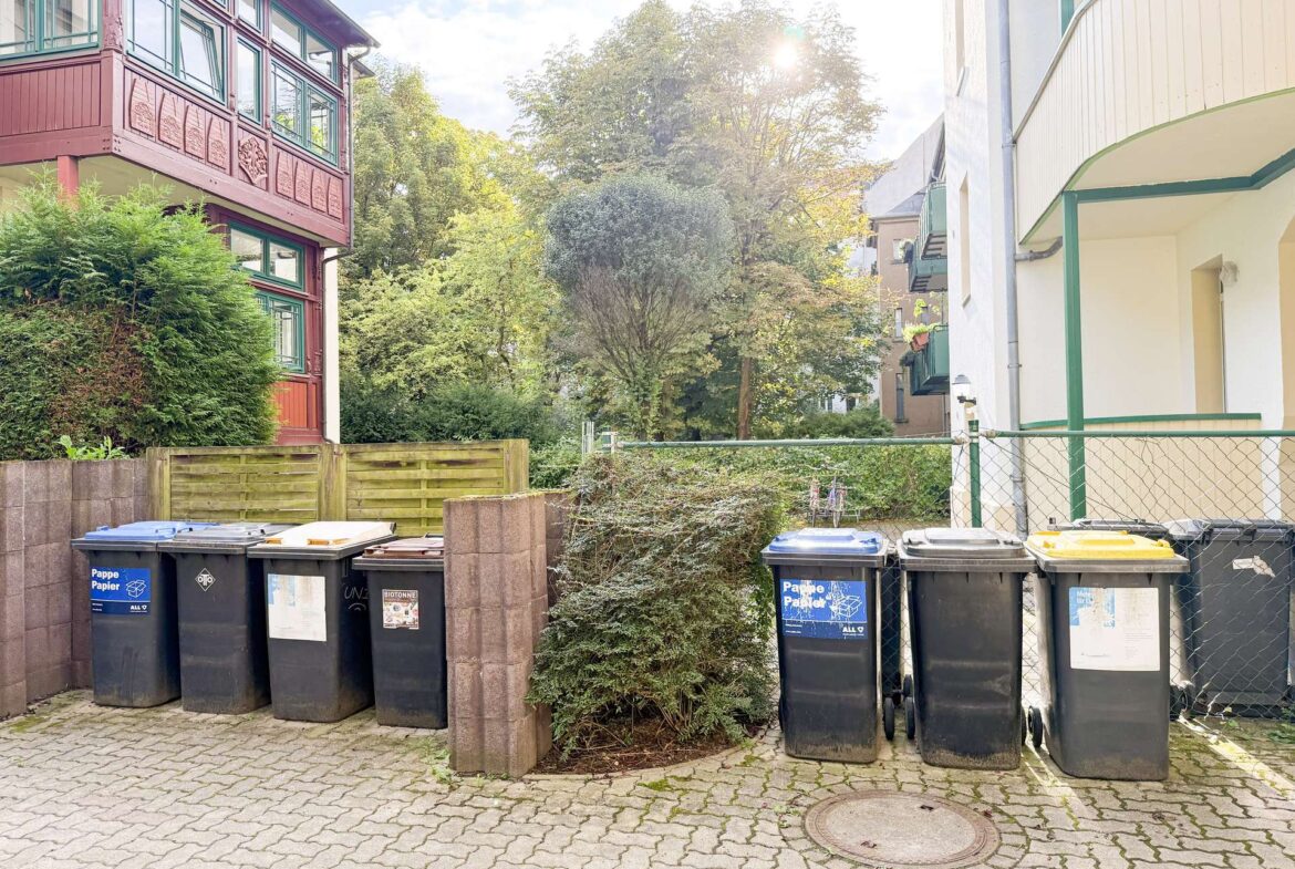 Row of black and blue recycling bins along a paved courtyard, flanked by red and white buildings and greenery behind them.