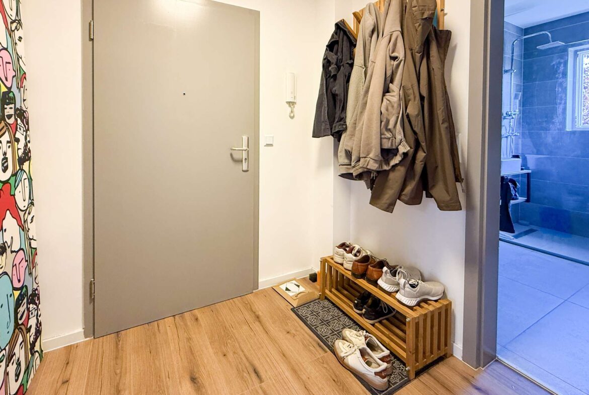 Entryway with a closed grey door, coat rack of jackets to the right, and a wooden shoe rack with sneakers along the wall; colorful mural on the left and a glass door leading to a blue-tiled bathroom on the right.