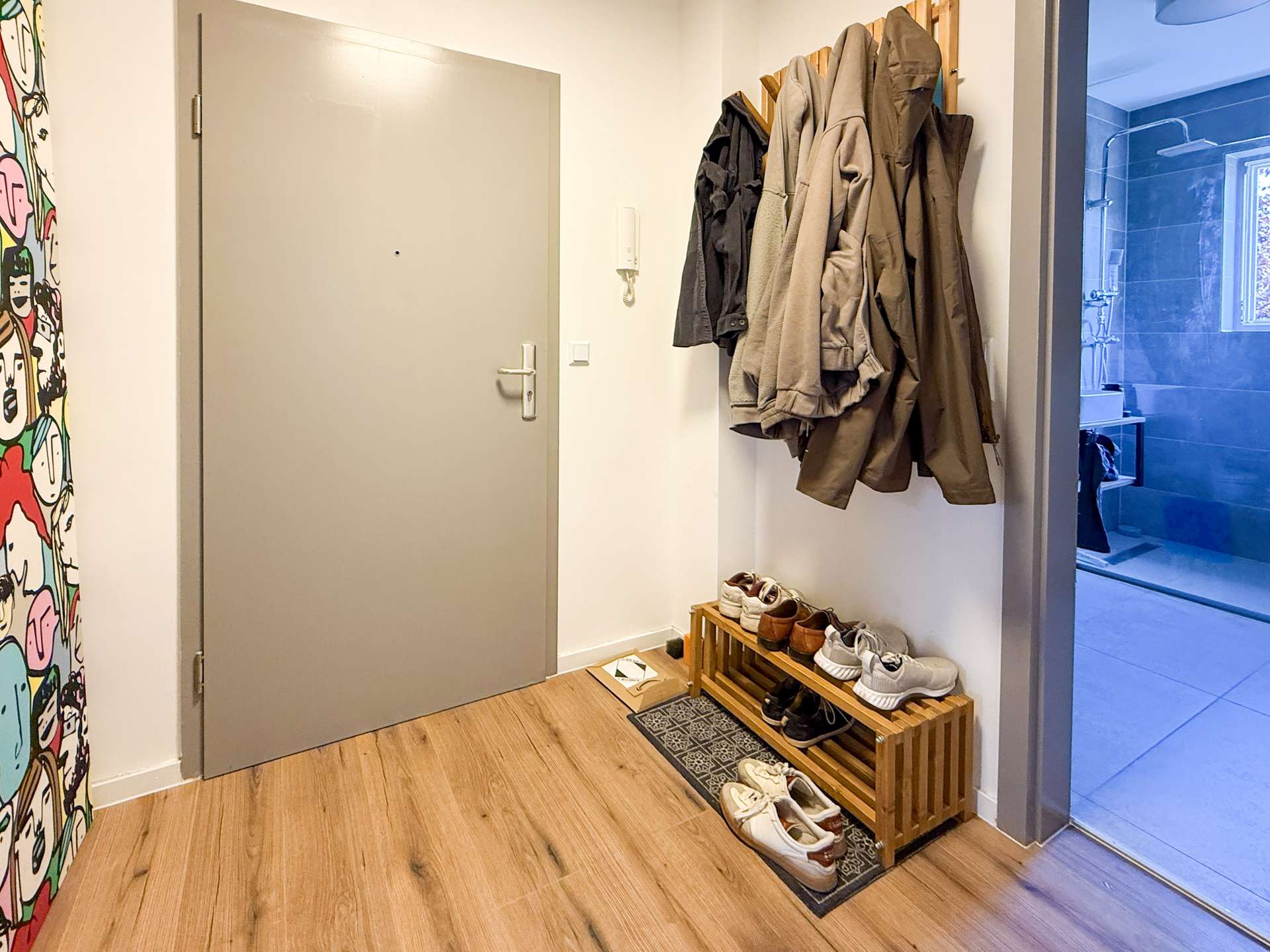 Entryway with a closed grey door, coat rack of jackets to the right, and a wooden shoe rack with sneakers along the wall; colorful mural on the left and a glass door leading to a blue-tiled bathroom on the right.
