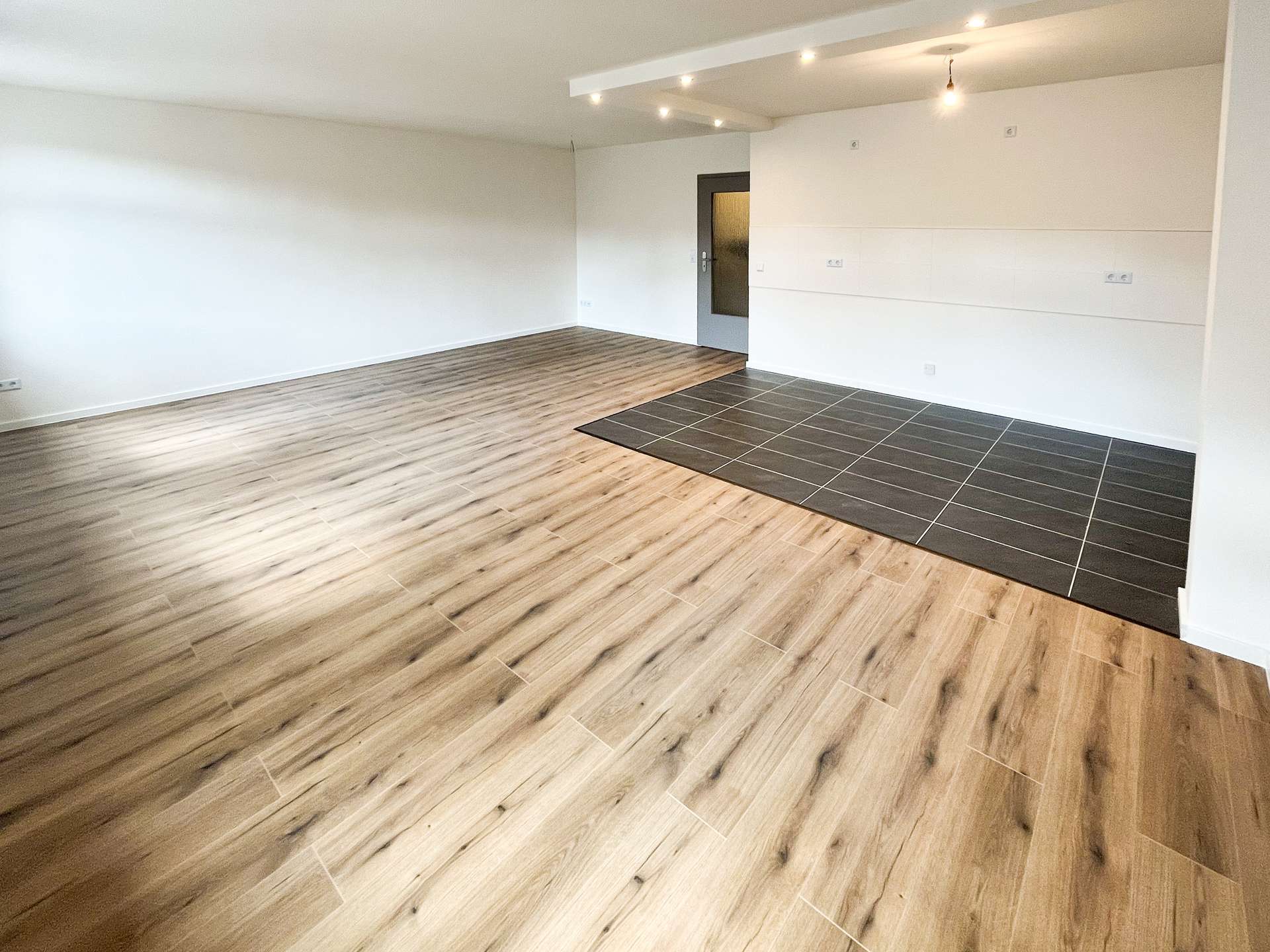 Bright, empty living area with light wood-look flooring, white walls, and a dark tile section near the kitchen area; gray door at the back wall; exposed ceiling lights create a warm ambiance.