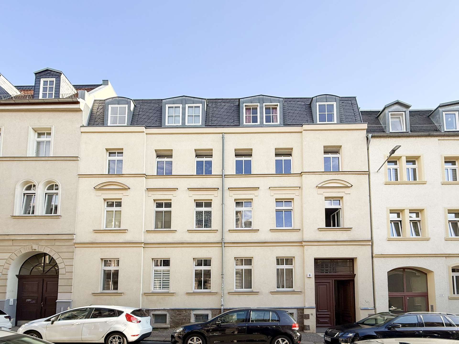 Row of light beige townhouses with dormer windows under a clear blue sky; cars parked along the curb in front of them.