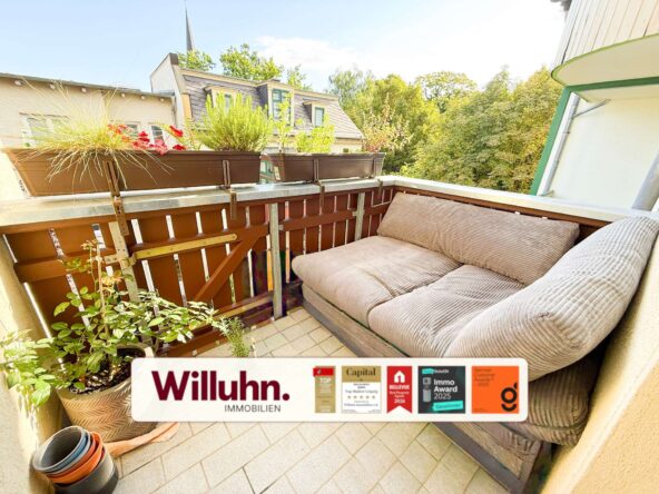 Cozy balcony with beige cushioned seating, potted plants along the railing, and a Willuhn Immobilien banner at the bottom.