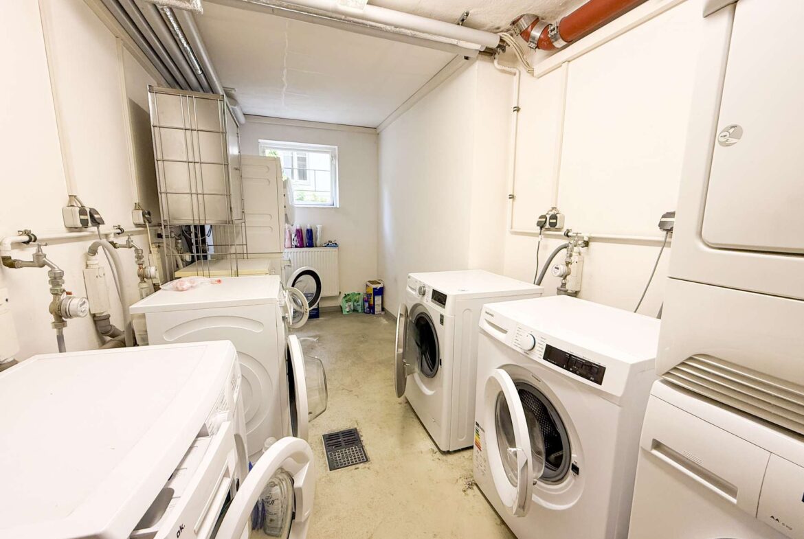 Narrow laundry room with multiple front-loading washing machines, exposed pipes along the ceiling, and a small window at the far end.