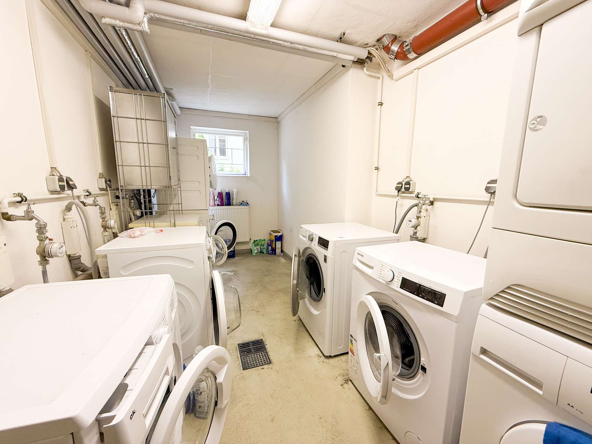 Narrow laundry room with multiple front-loading washing machines, exposed pipes along the ceiling, and a small window at the far end.