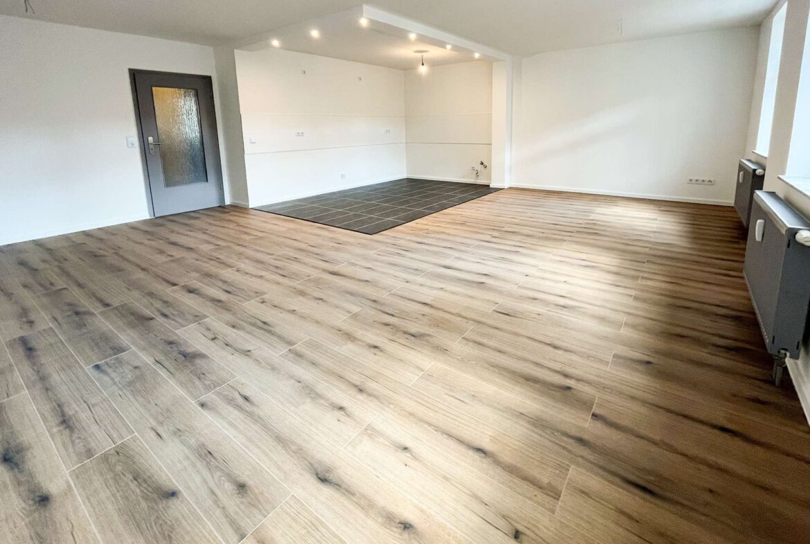 Empty open-concept living area with light wood-look flooring, white walls, a gray door, and a small dark-tiled section likely for a kitchen area in the back corner.