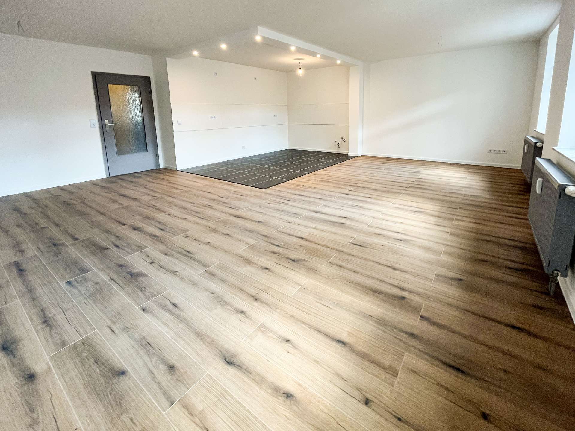 Empty open-concept living area with light wood-look flooring, white walls, a gray door, and a small dark-tiled section likely for a kitchen area in the back corner.