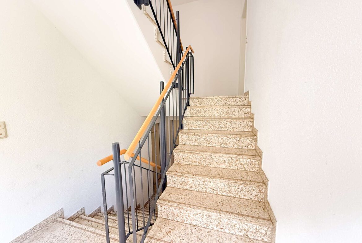 Interior staircase with terrazzo steps, black metal railing, and a light wooden handrail along white walls.