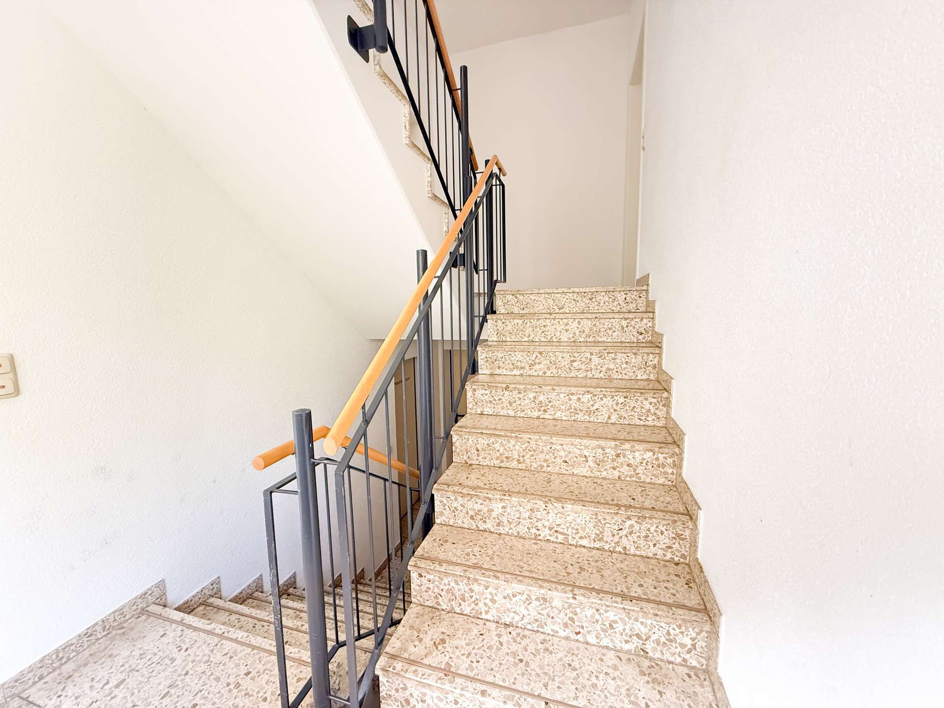 Interior staircase with terrazzo steps, black metal railing, and a light wooden handrail along white walls.