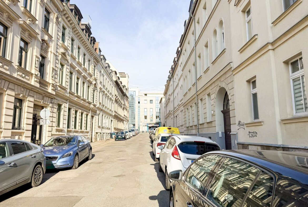 Narrow urban street flanked by parked cars and ornate beige buildings on the left, white buildings on the right, under a blue sky.