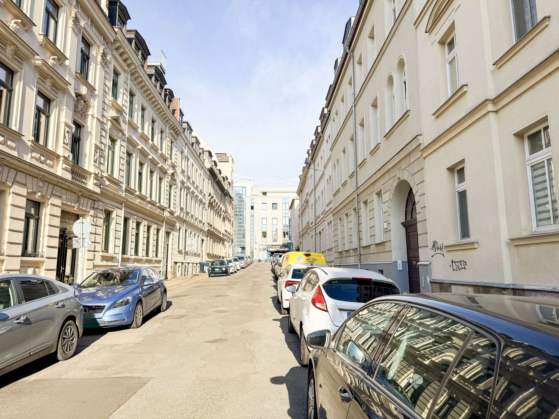 Narrow urban street flanked by parked cars and ornate beige buildings on the left, white buildings on the right, under a blue sky.