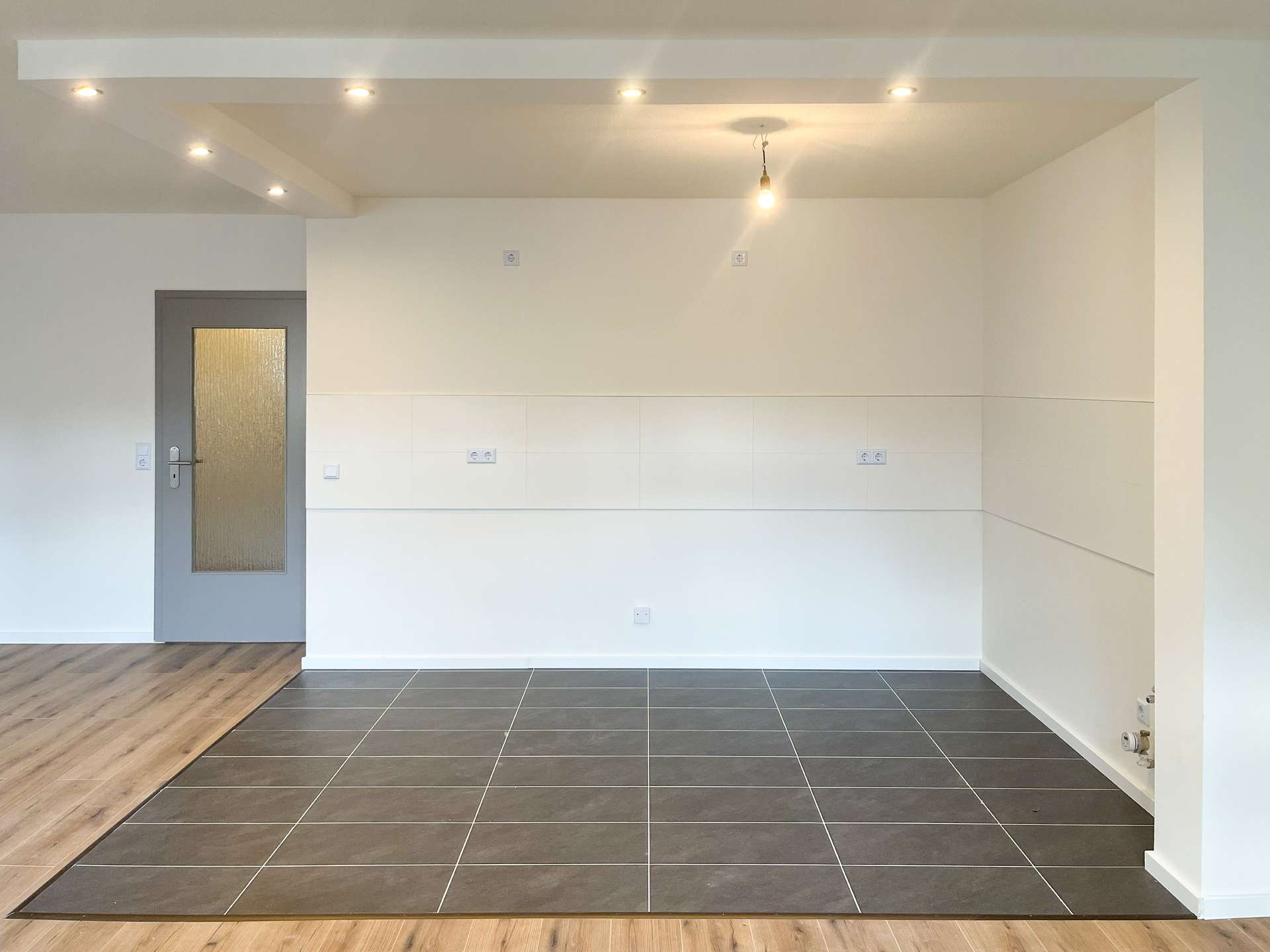 Empty modern interior with wood flooring along a step, dark tiled area, white walls, a gray door with frosted glass, and a hanging bulb.