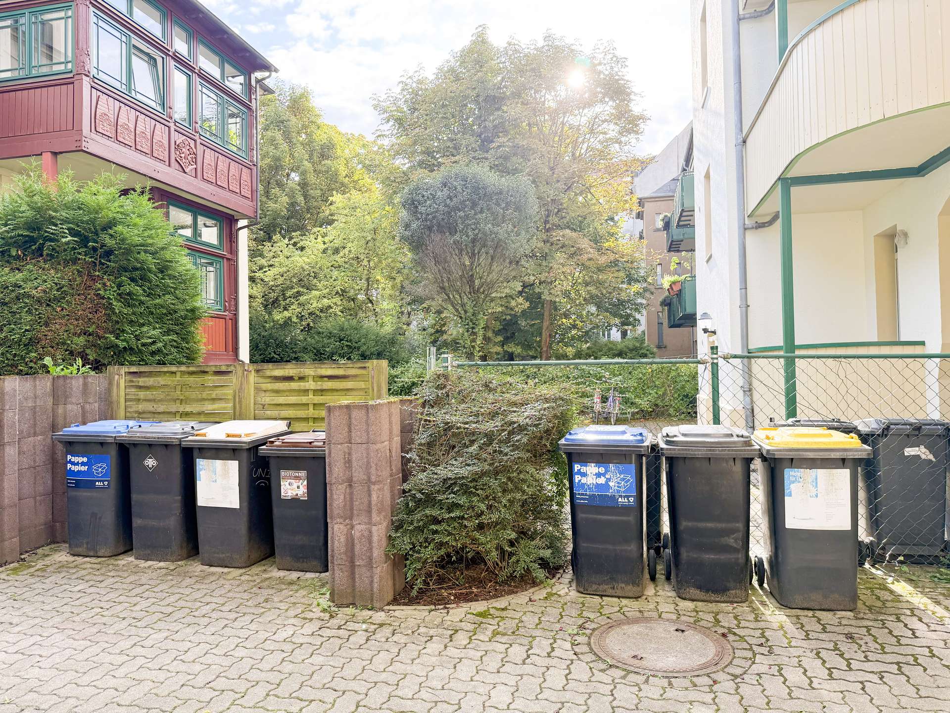 Row of dark blue and black recycling bins lined up along a brick-paved courtyard between red and white apartment buildings, with trees in the background