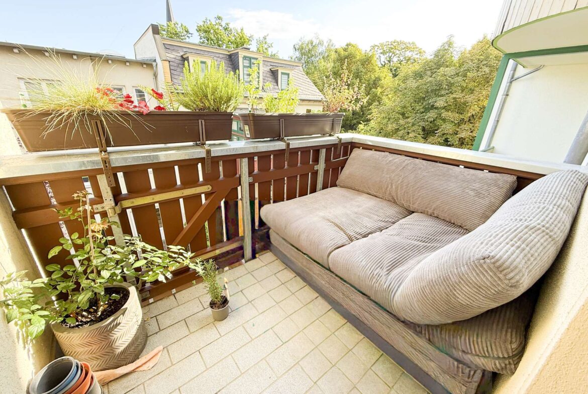 Cozy balcony seating area with beige cushioned sofa along a wooden railing, plus several potted plants in planters along the railing and floor tiles beneath, overlooking trees and neighboring rooftops.