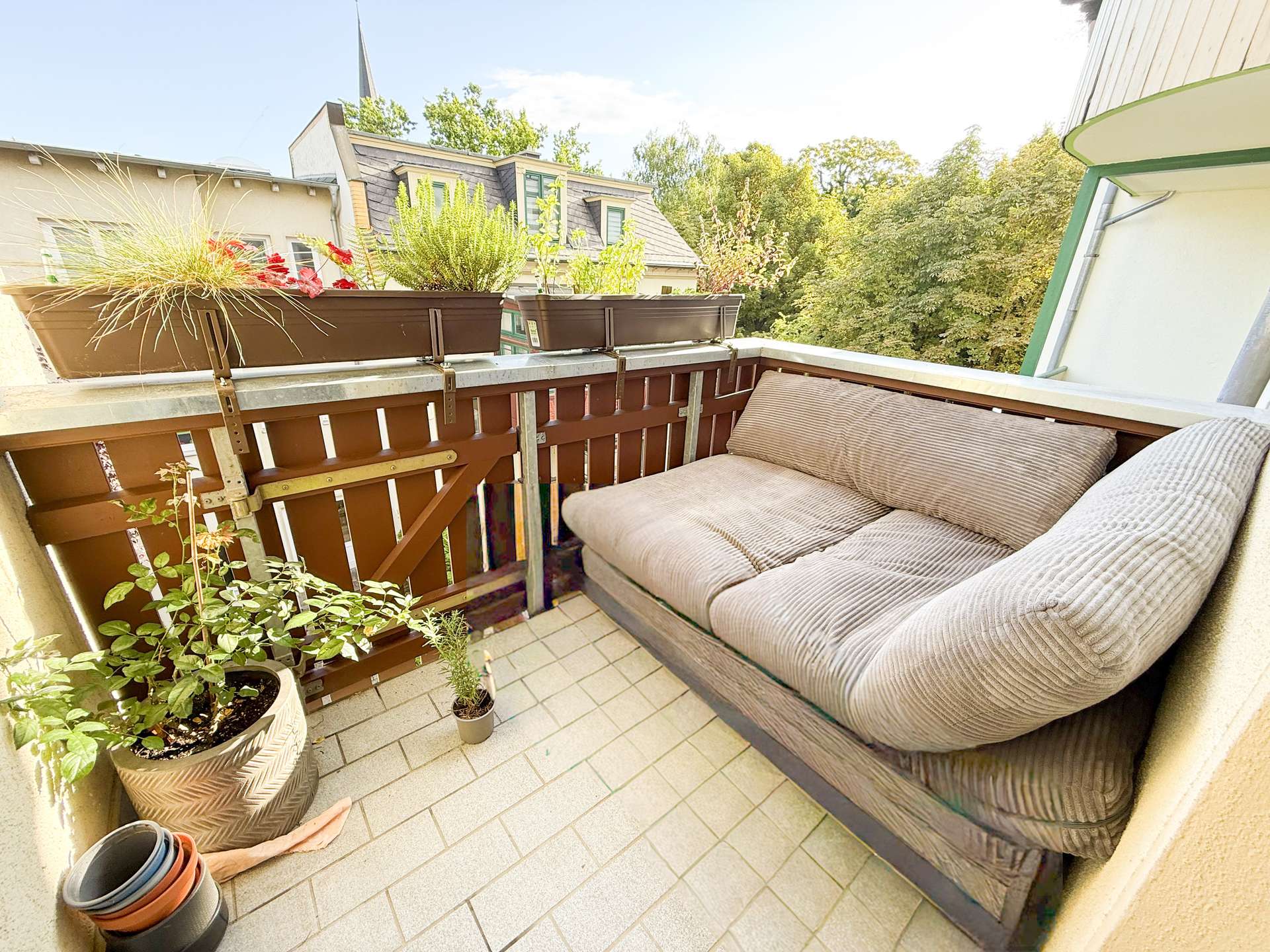 Cozy balcony seating area with beige cushioned sofa along a wooden railing, plus several potted plants in planters along the railing and floor tiles beneath, overlooking trees and neighboring rooftops.