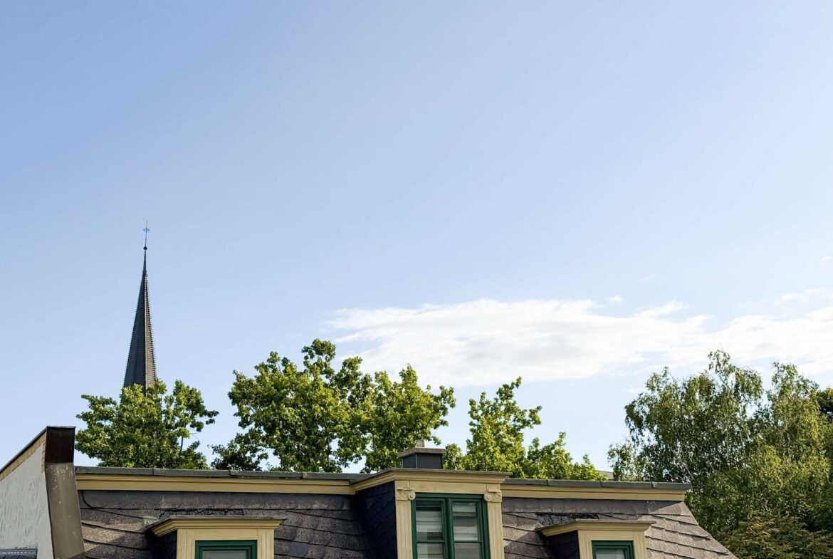 Slate-roofed building with dormer windows, green trim, and a church spire behind trees against a clear blue sky at the top left.