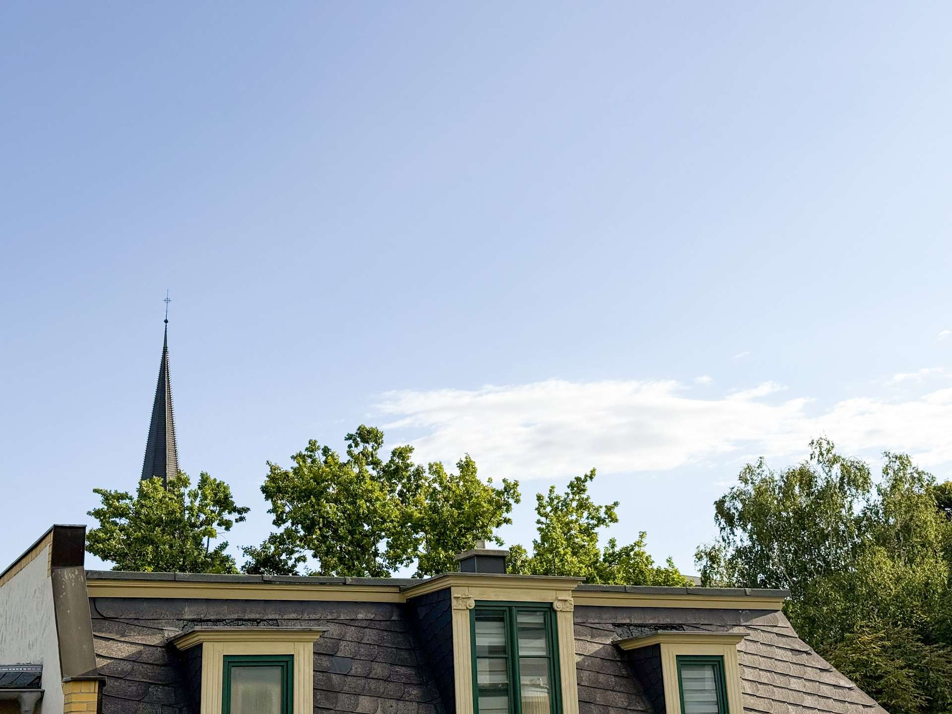 Slate-roofed building with dormer windows, green trim, and a church spire behind trees against a clear blue sky at the top left.