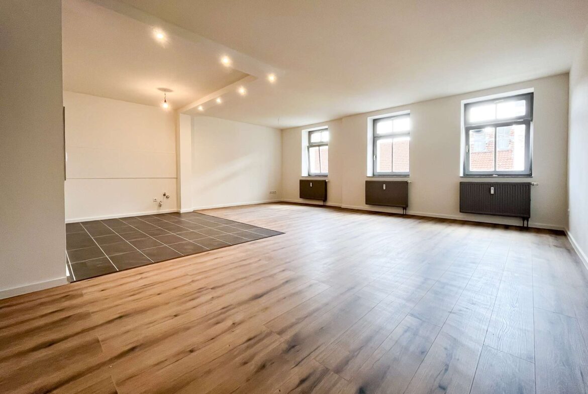 Bright empty living space with three windows along one wall, radiators beneath, and light wood flooring; a dark tiled area sits in the foreground.