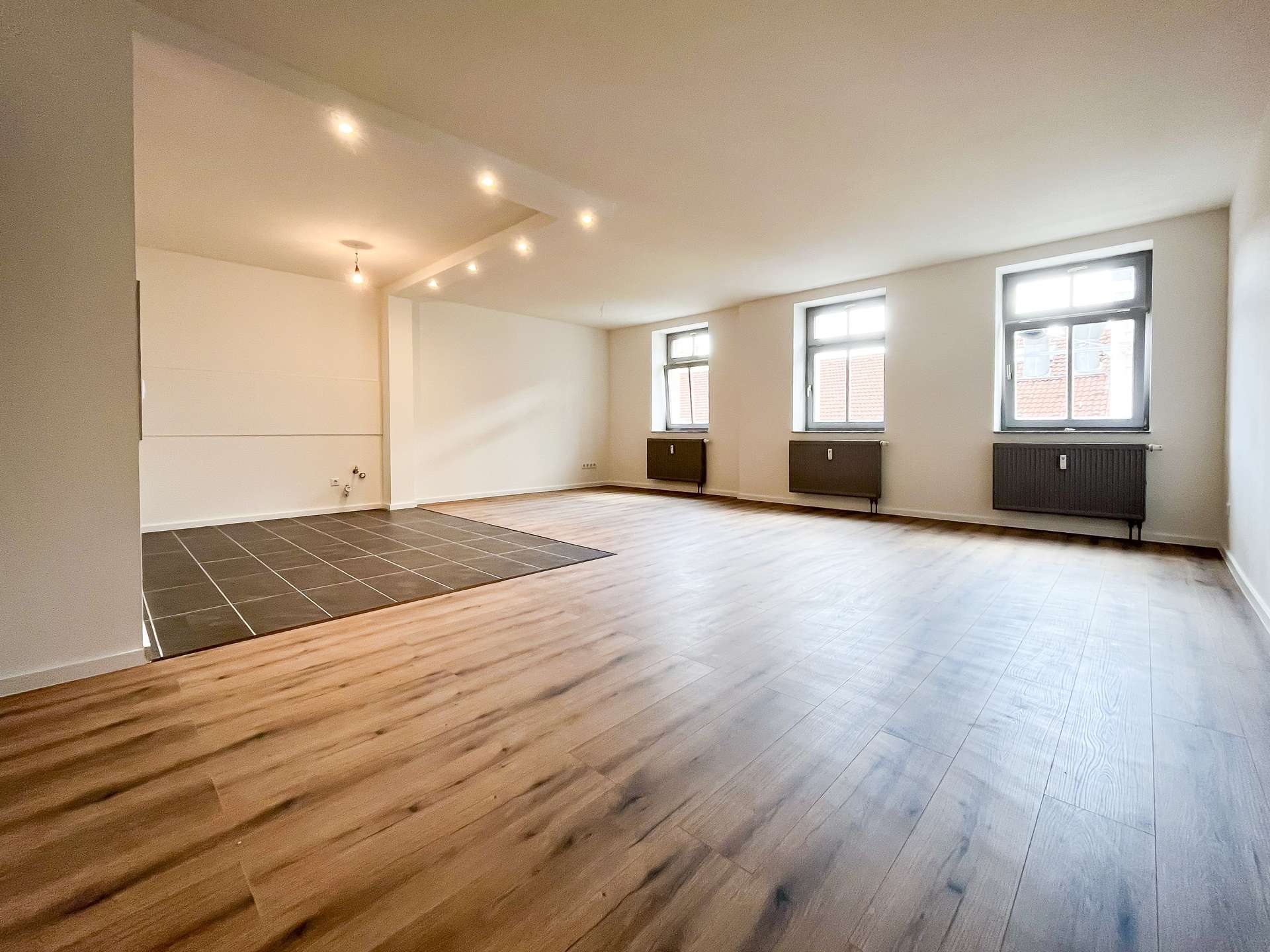 Bright empty living space with three windows along one wall, radiators beneath, and light wood flooring; a dark tiled area sits in the foreground.
