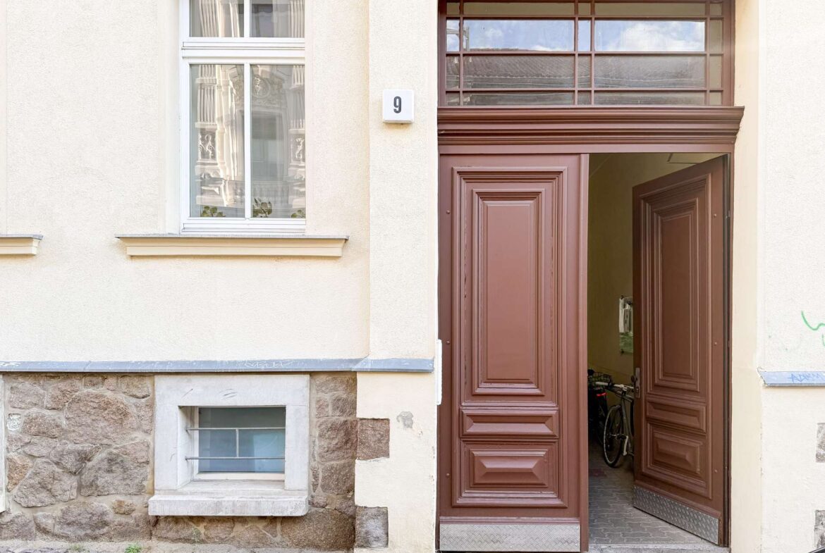 Beige building façade with a tall ornate brown double door open to a hallway; a white-framed window with curtains on the left; number 9 on the wall.