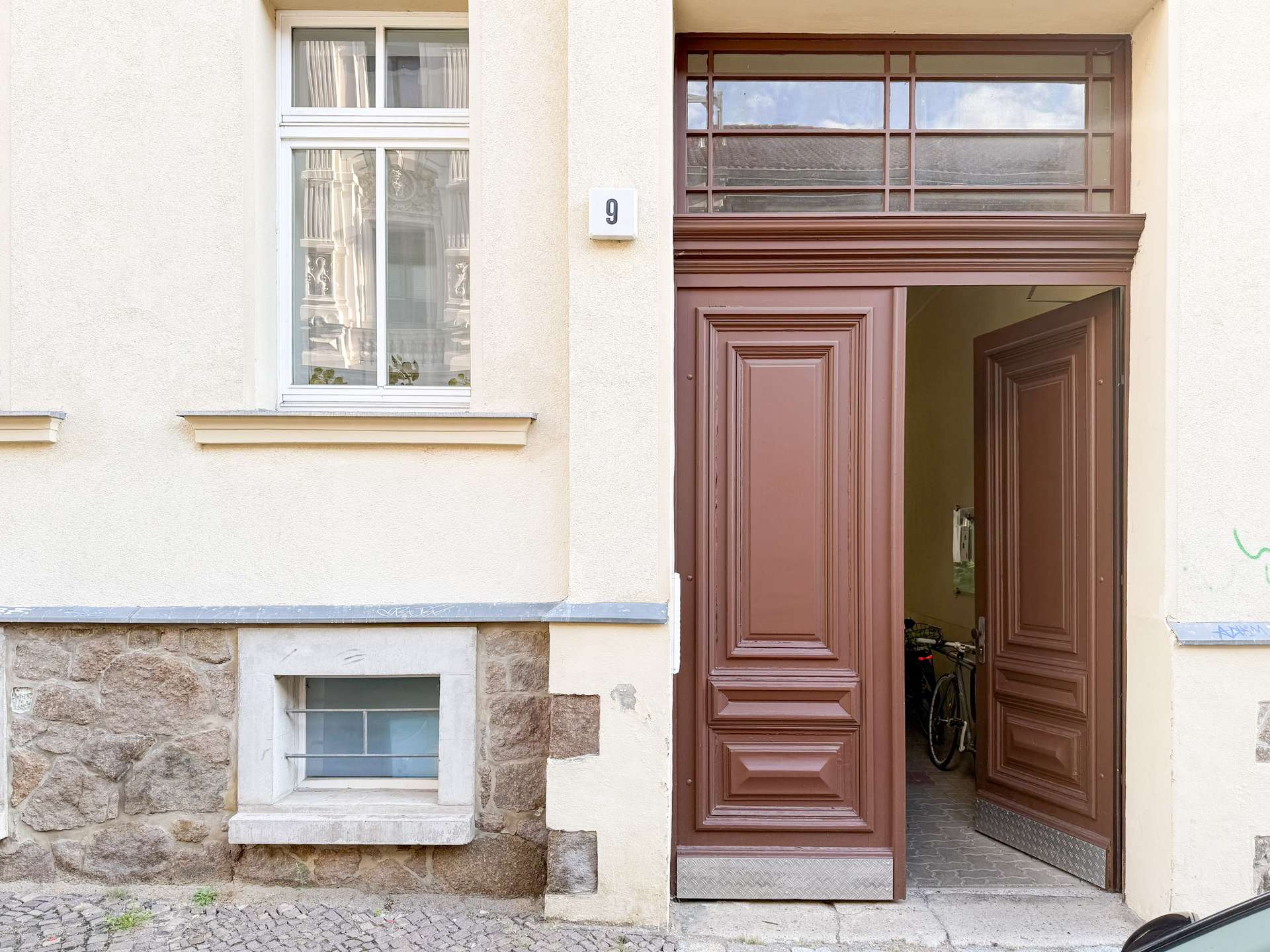 Beige building façade with a tall ornate brown double door open to a hallway; a white-framed window with curtains on the left; number 9 on the wall.
