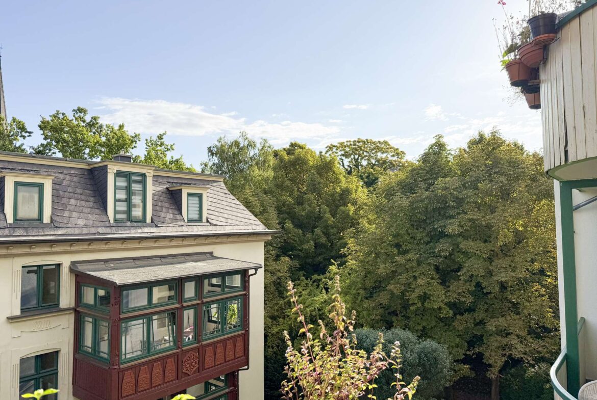 View from a balcony showing a beige building with green-framed windows and a decorative brown bay window, overlooking lush trees under a clear blue sky with a right-side balcony railing and potted plants.