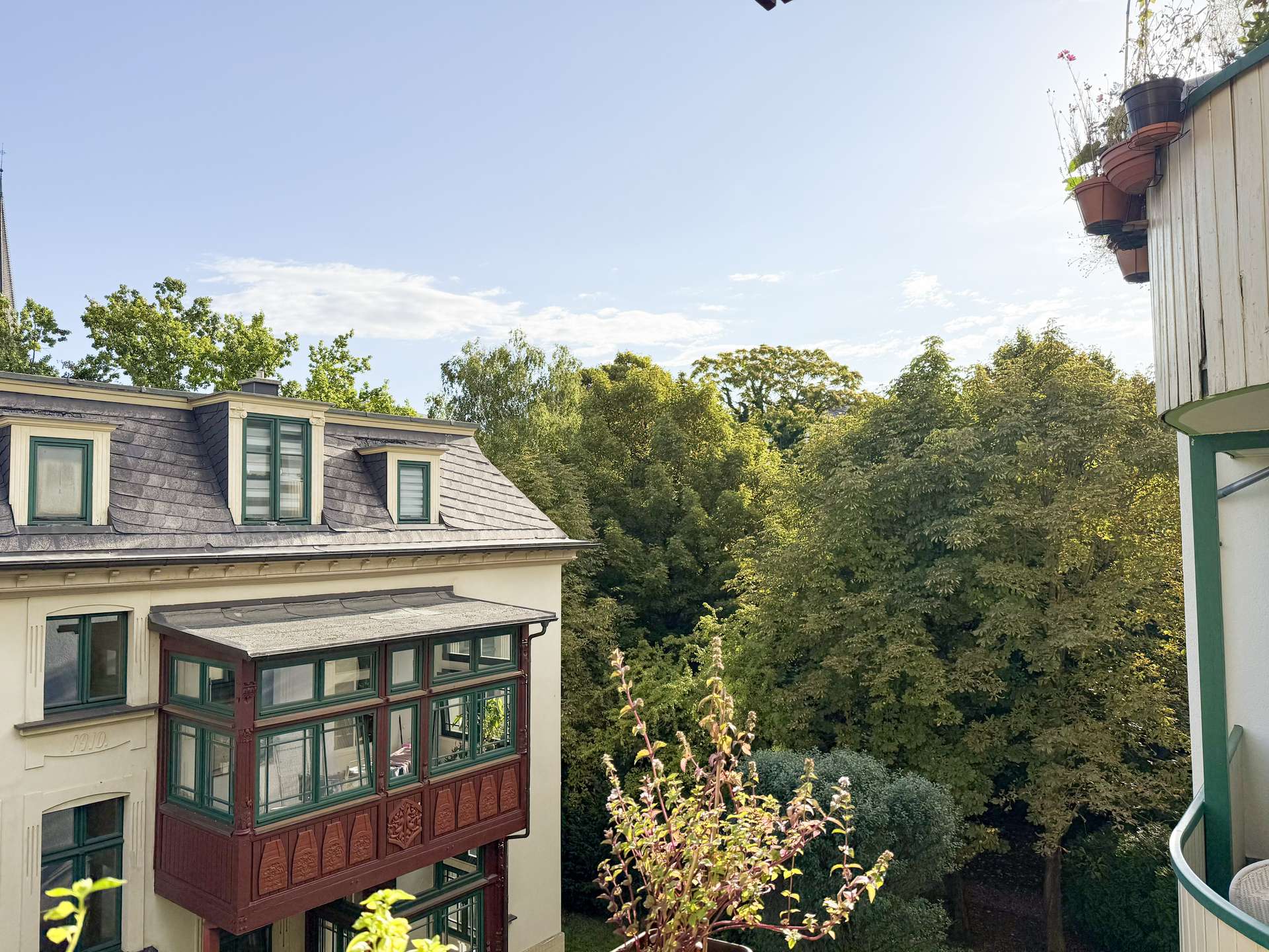 View from a balcony showing a beige building with green-framed windows and a decorative brown bay window, overlooking lush trees under a clear blue sky with a right-side balcony railing and potted plants.