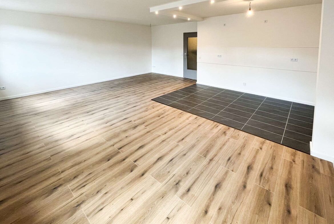 Empty open-plan room with light wood flooring and a dark tiled area near the wall, white walls, and a doorway.
