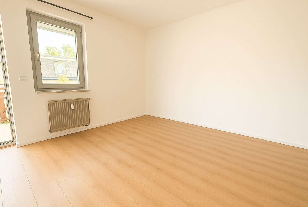 Bright empty room with light wood floor, white walls, a radiator under a window, and a door leading to outside.