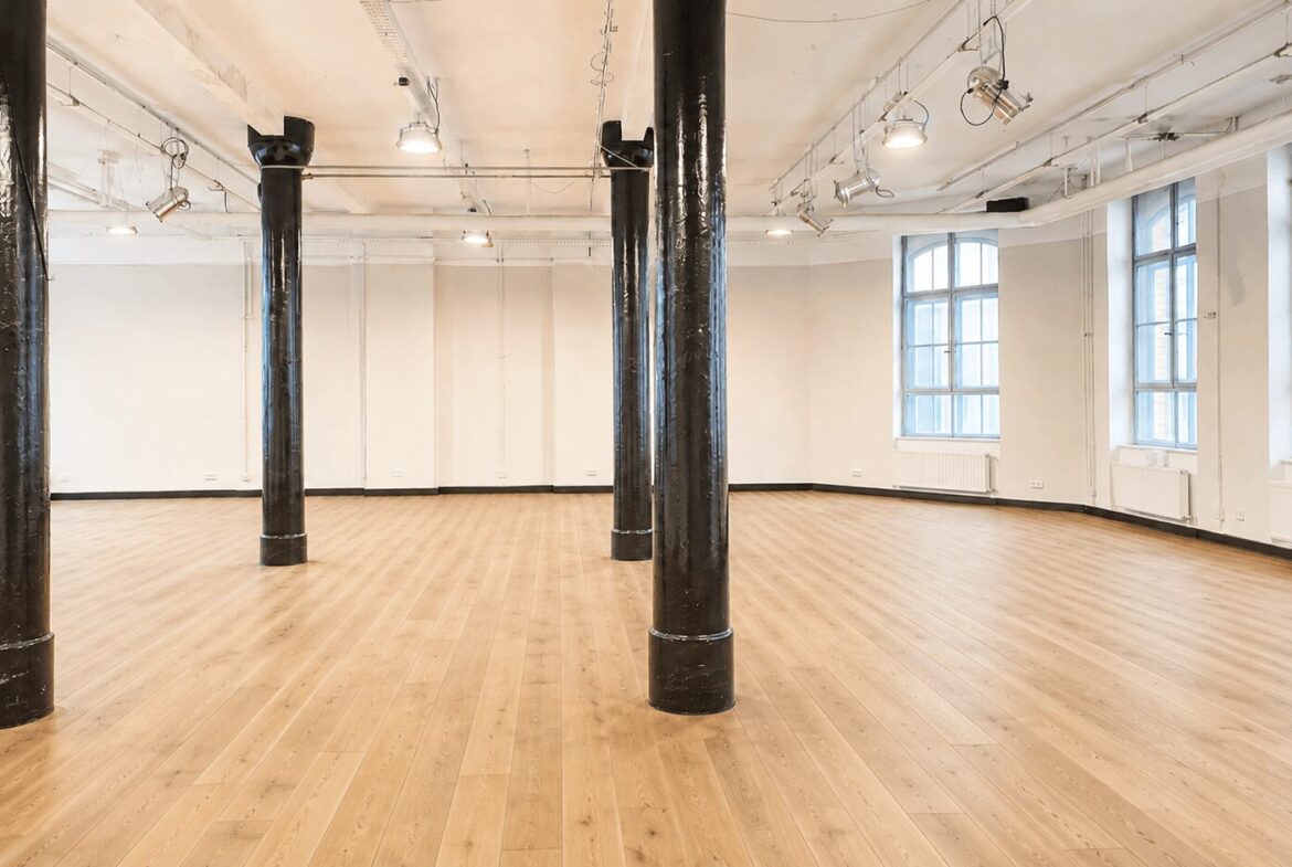 Empty, well-lit studio with a light wood floor, white walls, large windows, and black support pillars.