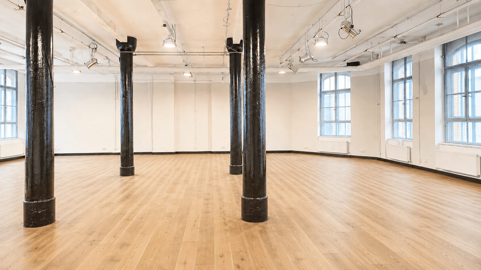 Empty, well-lit studio with a light wood floor, white walls, large windows, and black support pillars.