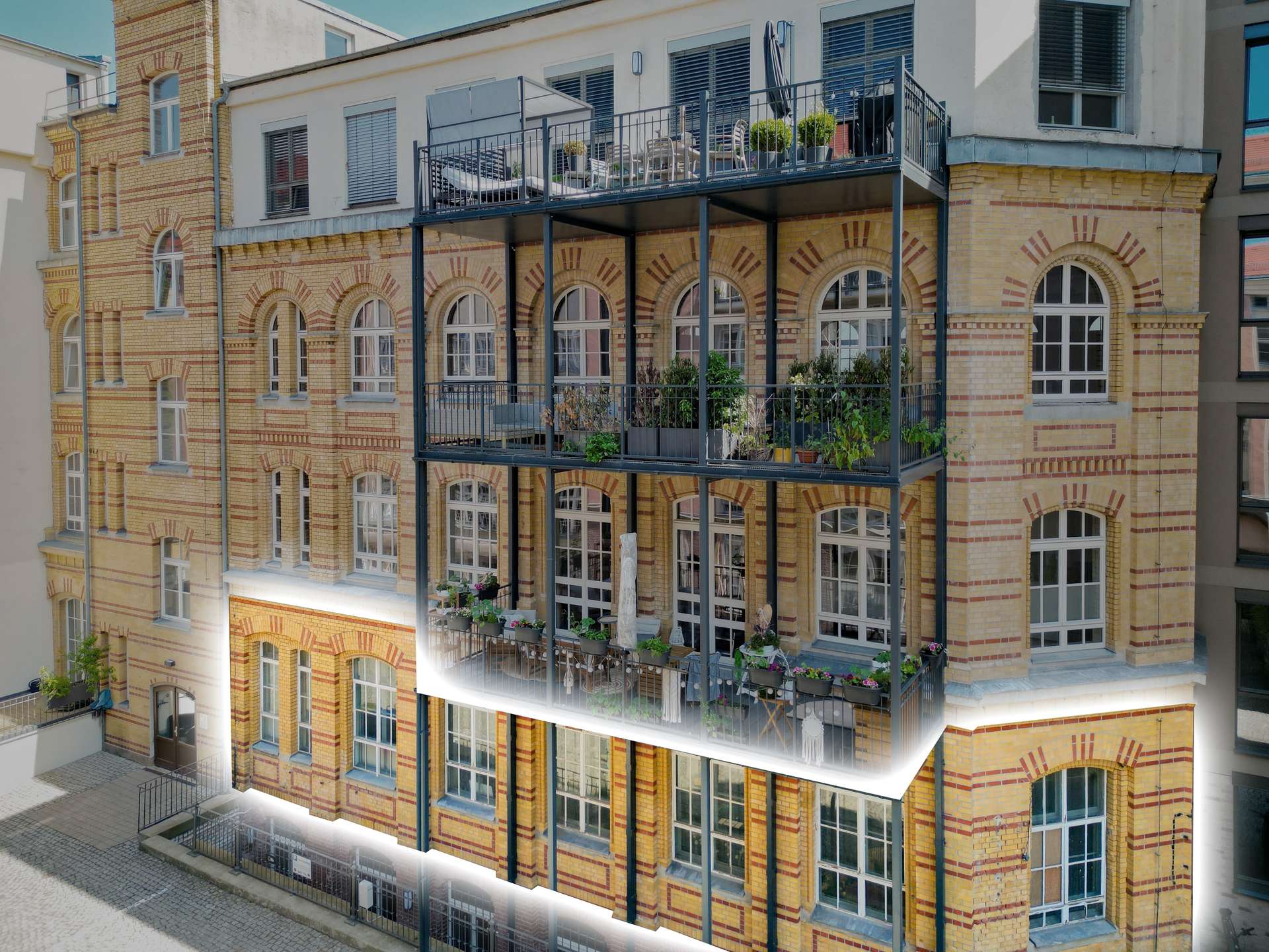 Brick apartment building with arched windows and three-tier metal balconies loaded with potted plants and furniture on each level, urban setting