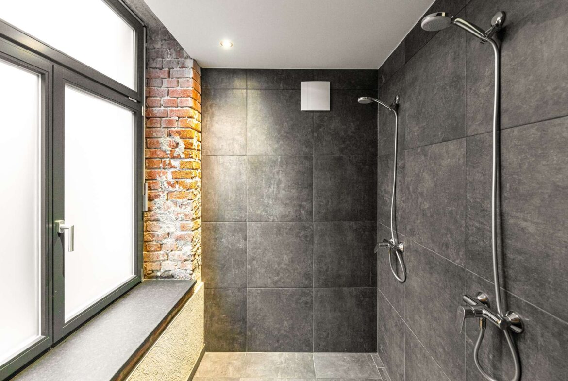 Walk-in shower with dark gray tile walls, an exposed brick pillar, a large window to the left, and chrome shower fixtures.