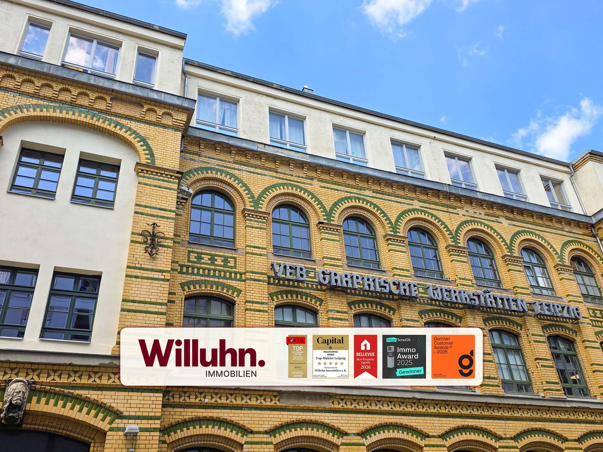 Brick building with decorative green arches and large arched windows, featuring a 'Willuhn Immobilien' banner and award plaques on the storefront.