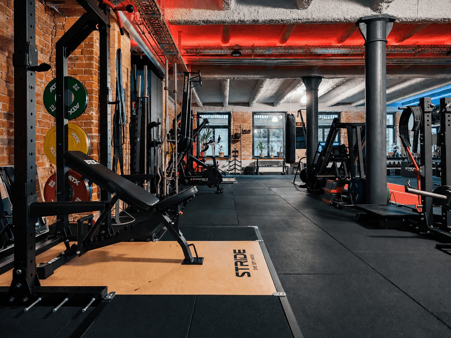 Industrial-style gym with weight racks, benches, and black rubber flooring; exposed brick walls and red lighting.