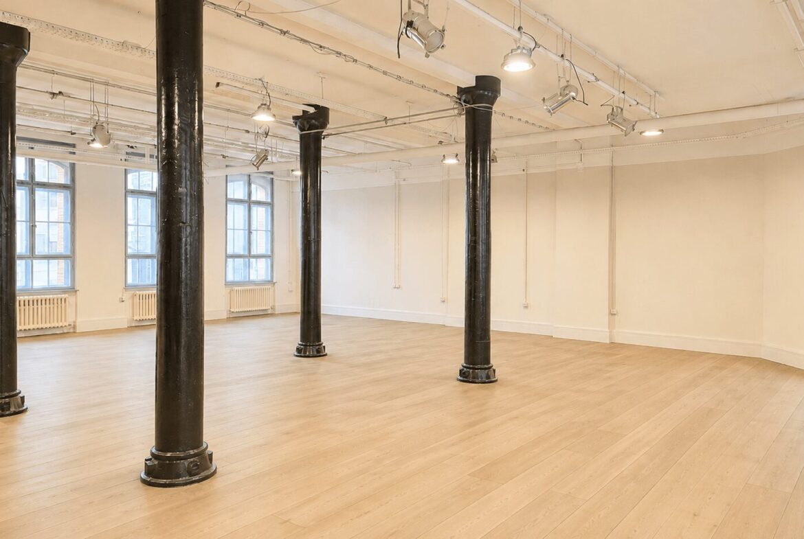 Empty industrial-style loft studio with four black support columns, light wood floor, and large arched windows letting in natural light